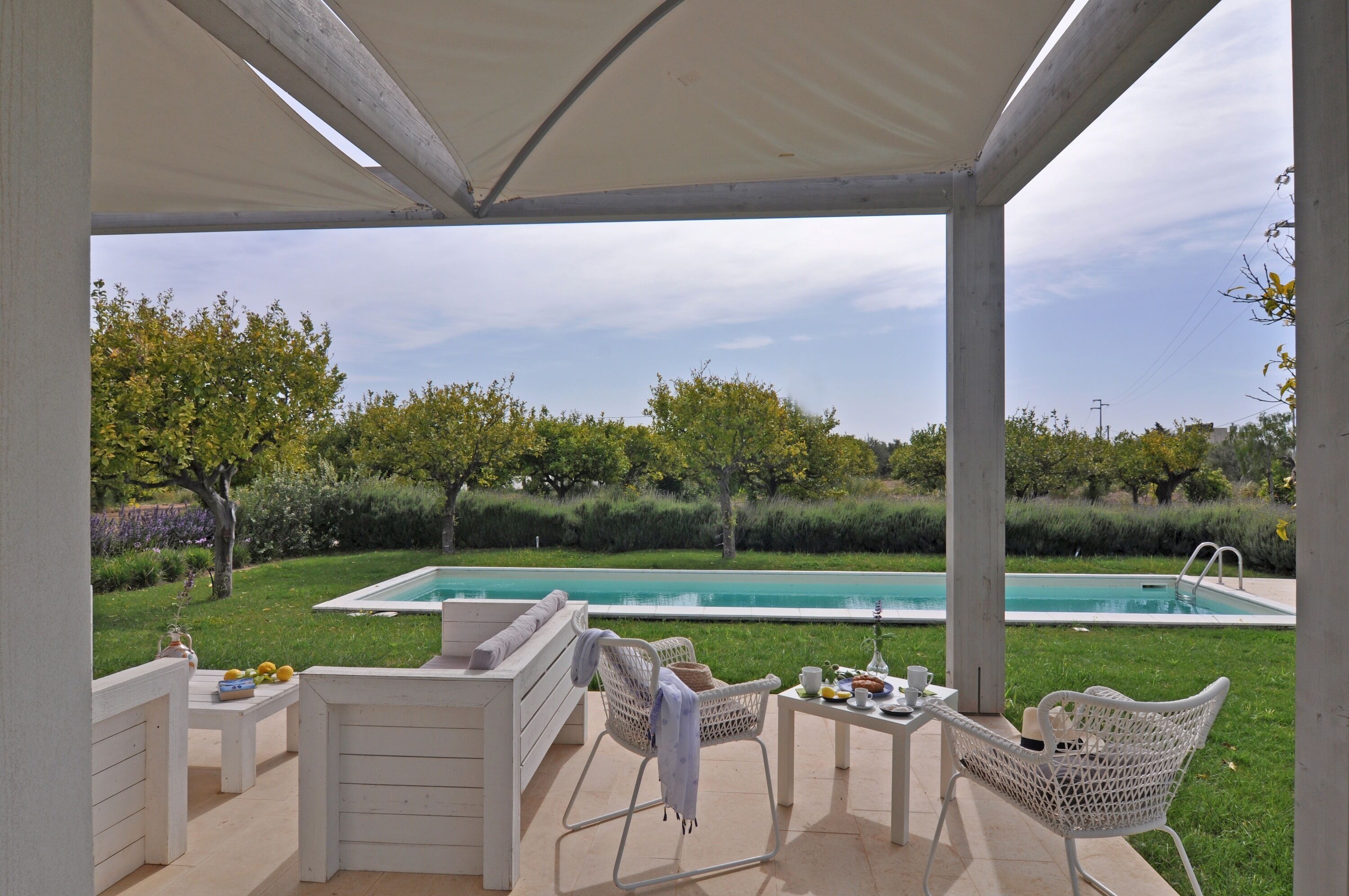 Un espace extérieur abrité, doté d’une piscine, de mobilier blanc et offrant une vue sur un jardin.