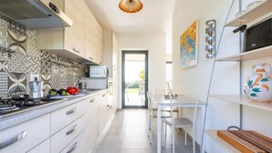 A modern kitchen with white cabinets, a gas stove, and a coffee maker. There is a dining area with a white table and chairs. - Villa Papeda in Noto (Noto)