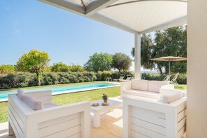 Un espacio al aire libre techado con zona de estar, piscina, muebles blancos y vistas a la piscina y follaje.