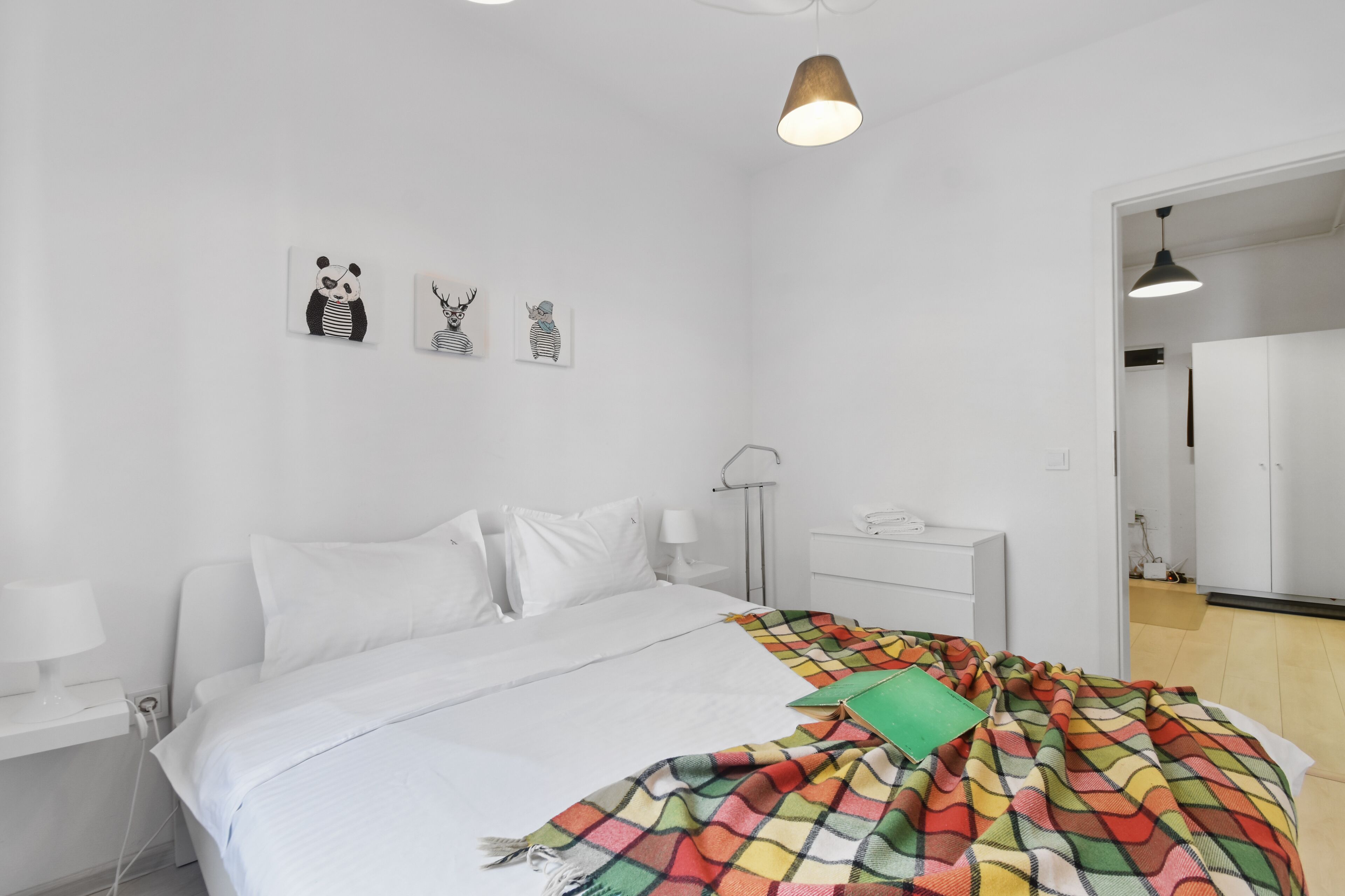 A bedroom with a bed, white walls, a bedside table with a lamp, a nightstand, and a door leading to another room.
