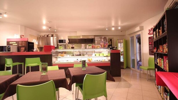 Un restaurant avec des chaises et des tables vertes, un comptoir où sont exposés les plats, et un espace cuisine.