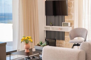 A modern living room with a flat-screen TV mounted on a stone wall, a comfortable grey armchair, a coffee table with a vase of yellow flowers, and a small shelf with decorative items.