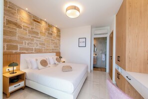 Un dormitorio moderno con una pared de piedra, una cama con ropa de cama blanca, una mesita de noche con una lámpara y armarios de madera.