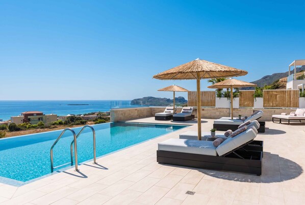 A pool area with lounge chairs and umbrellas, overlooking a clear blue sea.