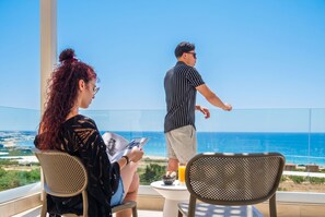 Un balcón con vistas despejadas al océano, una mesa con un vaso de jugo de naranja y dos personas sentadas.