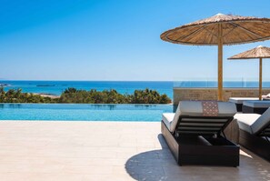 Zona de piscina con sillones reclinables y sombrillas, con vista al mar azul y transparente.