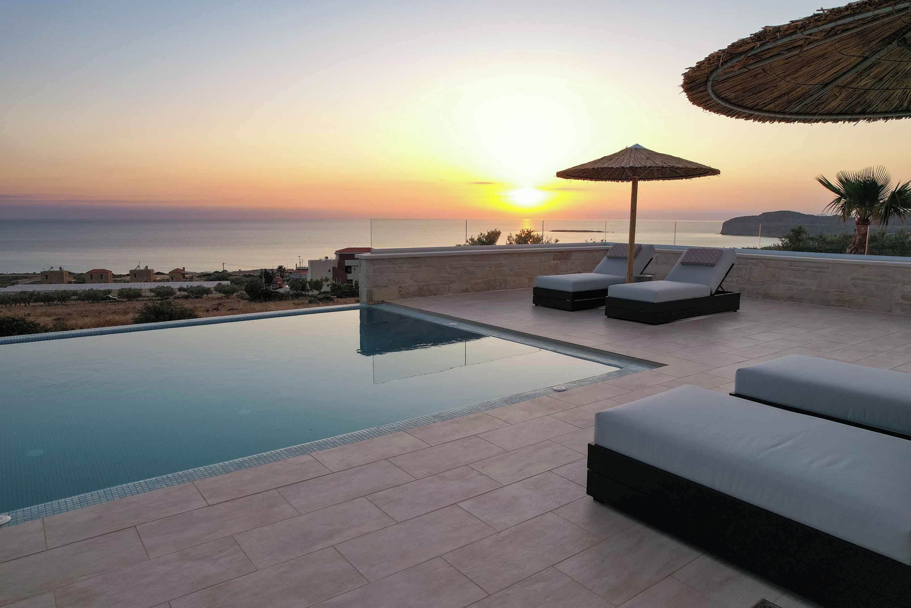 An infinity pool with lounge chairs and a thatched umbrella, overlooking a sunset over the sea.