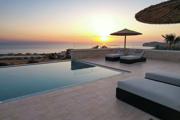 An infinity pool with lounge chairs and a thatched umbrella, overlooking a sunset over the sea.