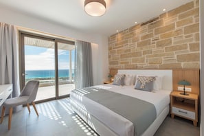 A bedroom with a large bed, a desk, and a chair. There is a view of the ocean through a sliding door.