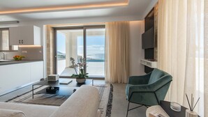 A modern hotel room with a large window offering a view of the sea, a comfortable seating area with a sofa and armchair, a flat-screen TV mounted on the wall, and a small kitchen area with a fruit bowl and potted plant. - Actea Seaview Villa 2 Free* heated pool & 800m sea (Kissamos)