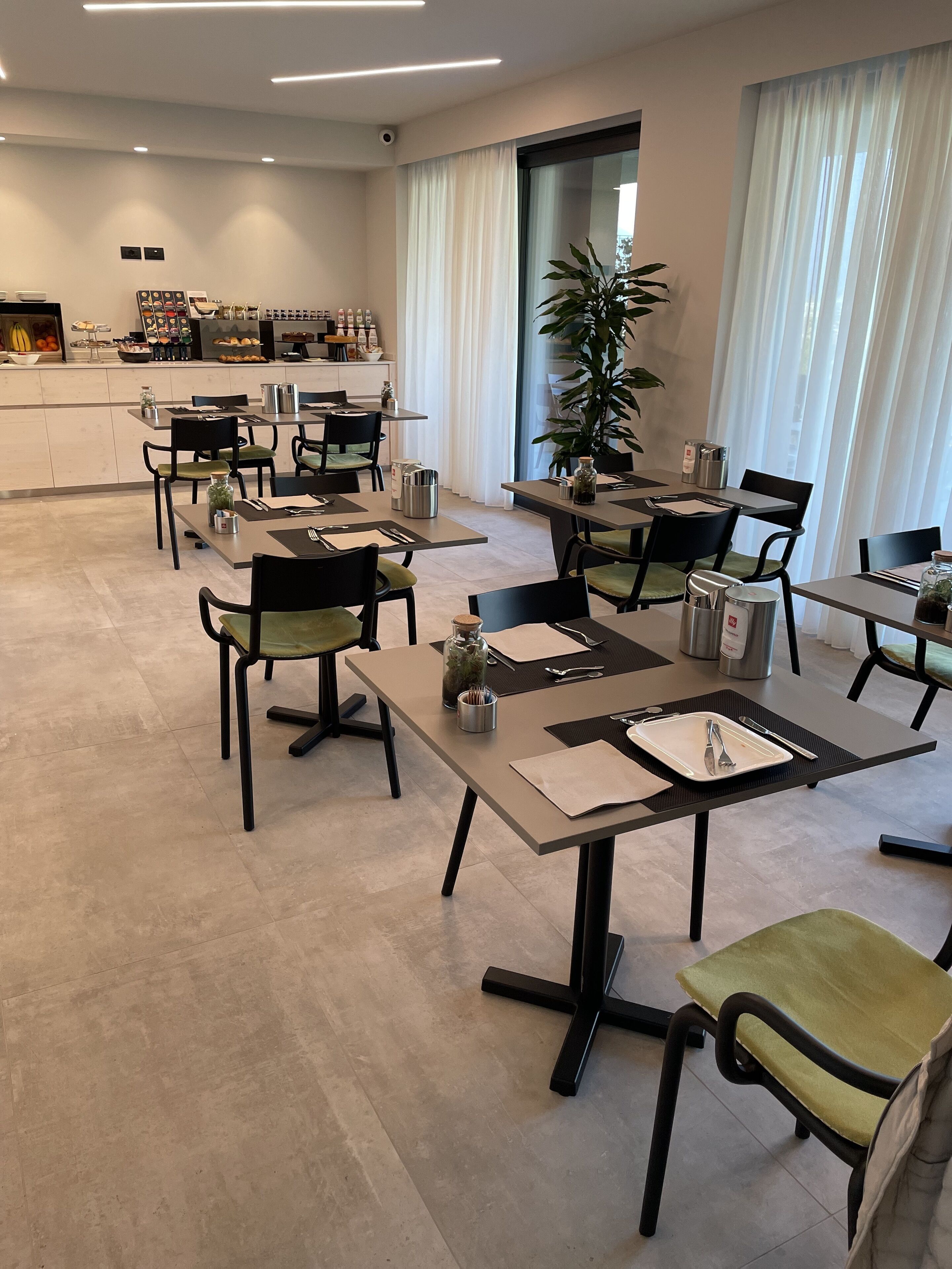 A modern dining area with tables and chairs, a kitchen in the background, and a large window with curtains.