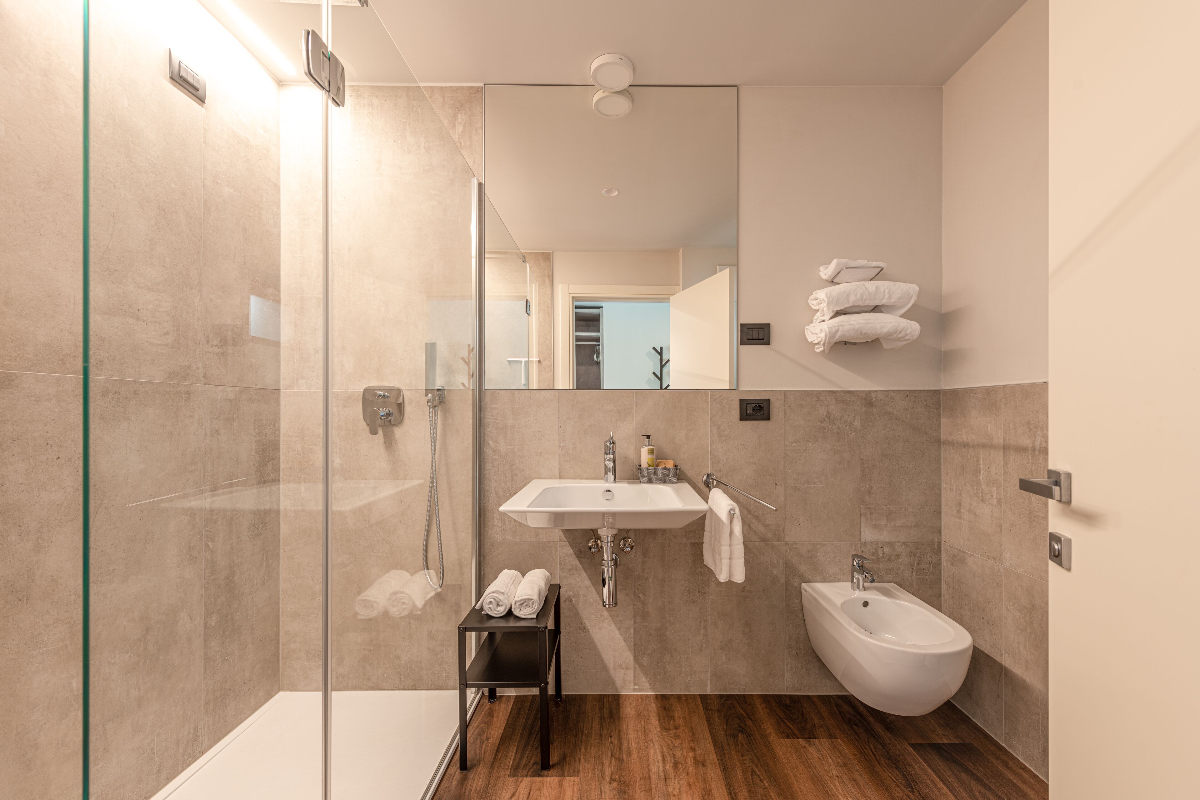 A modern bathroom with a glass shower enclosure, a sink, a bidet, and a small shelf with towels.