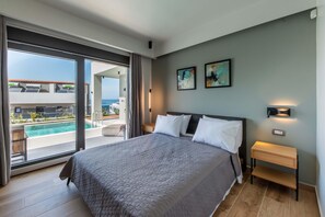 A modern bedroom with a large bed, a nightstand, and a view of a swimming pool and building. - Lusol Seafront Villas-Free* Heated Pools, 100m Sea (Chania)