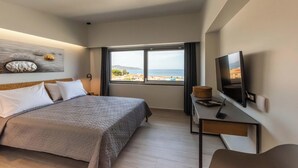 A modern hotel room with a large bed, a desk with a laptop, a flat-screen TV, and a window offering a view of the sea.