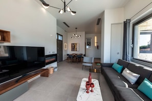 A modern living room with a flat-screen TV, a sofa, and a dining area with a table and chairs. - Lusol Seafront Villas-Free* Heated Pools, 100m Sea (Chania)
