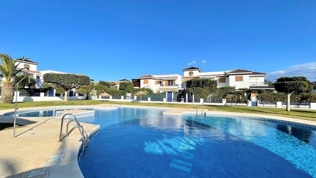 Een zwembad met een helderblauw wateroppervlak, omringd door een verzorgd gazon en een rij huizen in mediterrane stijl.