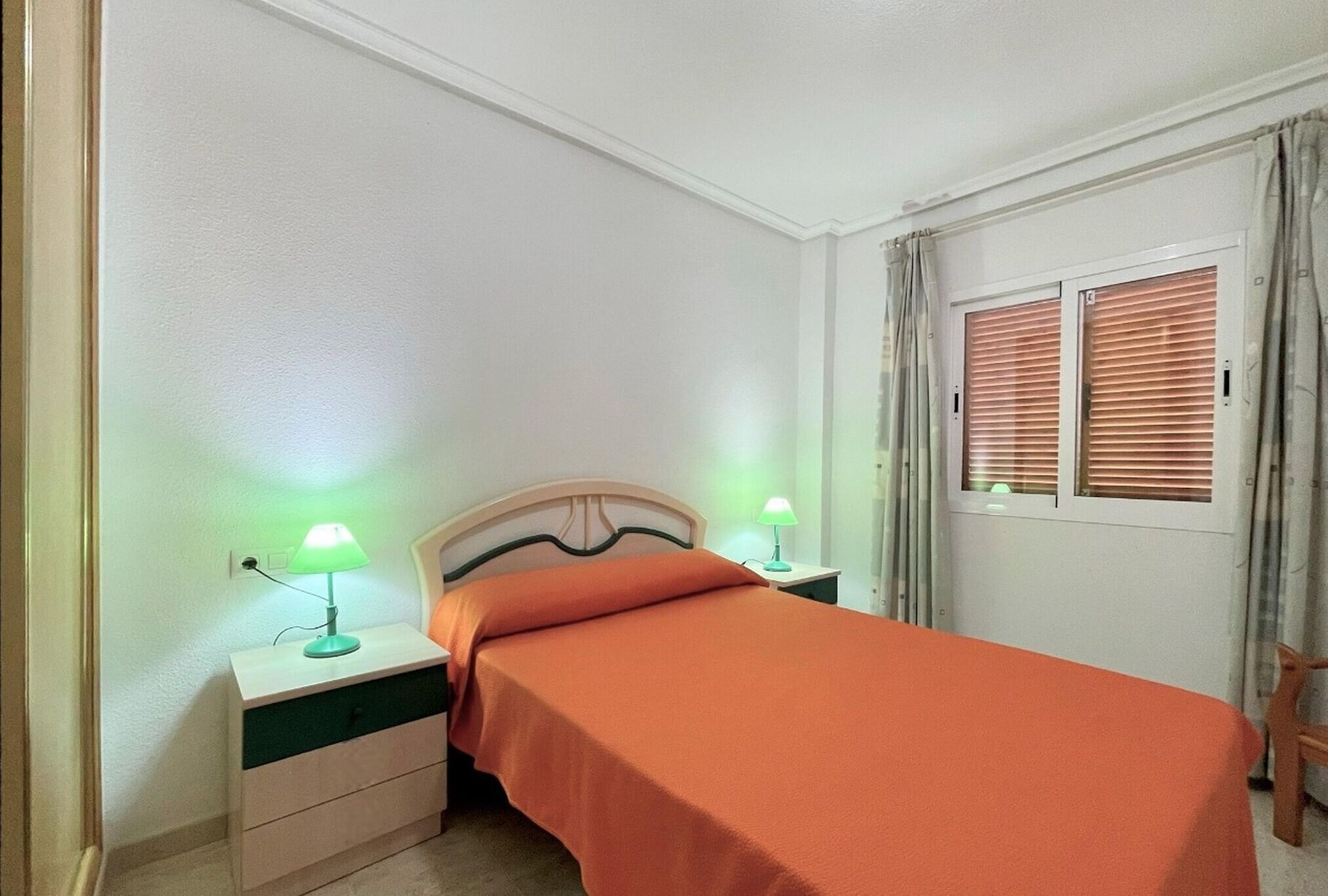 A bedroom with a bed, two bedside tables with lamps, a window with blinds, and a door.