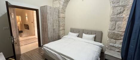 Une chambre avec une arche en pierre, un lit recouvert de draps blancs, une armoire en bois et une petite table avec une chaise.