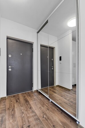 Una habitación con suelo de madera, una puerta oscura y un espejo grande que refleja la habitación.