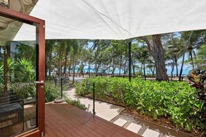 A wooden deck with a glass door leading to a shaded outdoor area with a view of the beach and palm trees. - Onda Beach | 4 Bedroom Home with Heated Pool (Clifton Beach)