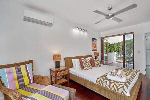 A bedroom with a large bed, a wicker chair, a nightstand with a lamp, and a ceiling fan. - Onda Beach | 4 Bedroom Home with Heated Pool (Clifton Beach)