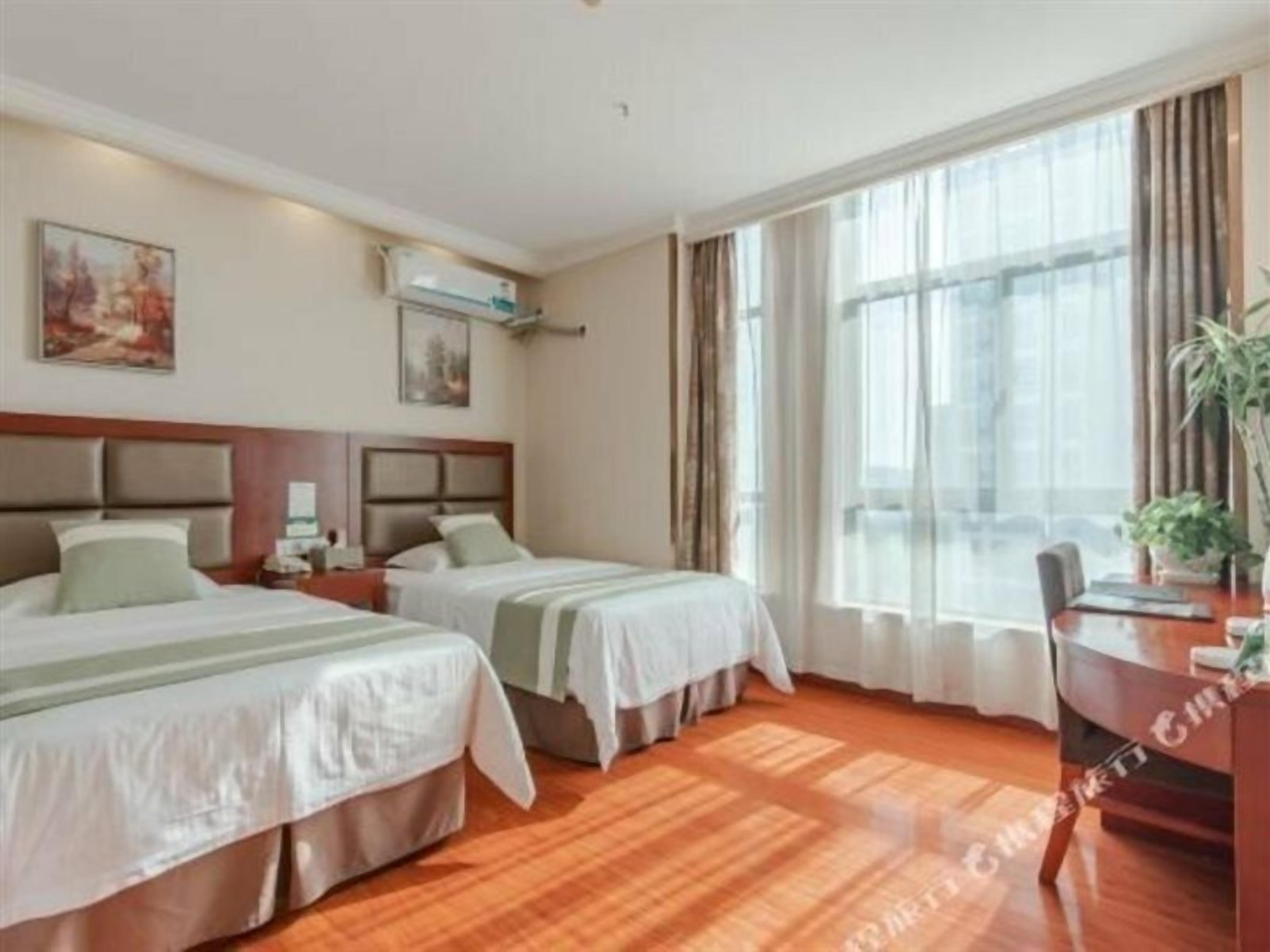 A hotel room with two beds, a wooden floor, a large window with curtains, and a desk with a chair.