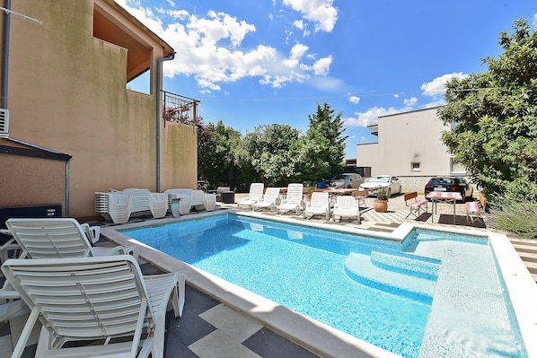 A swimming pool with lounge chairs and a covered area, surrounded by trees and buildings.