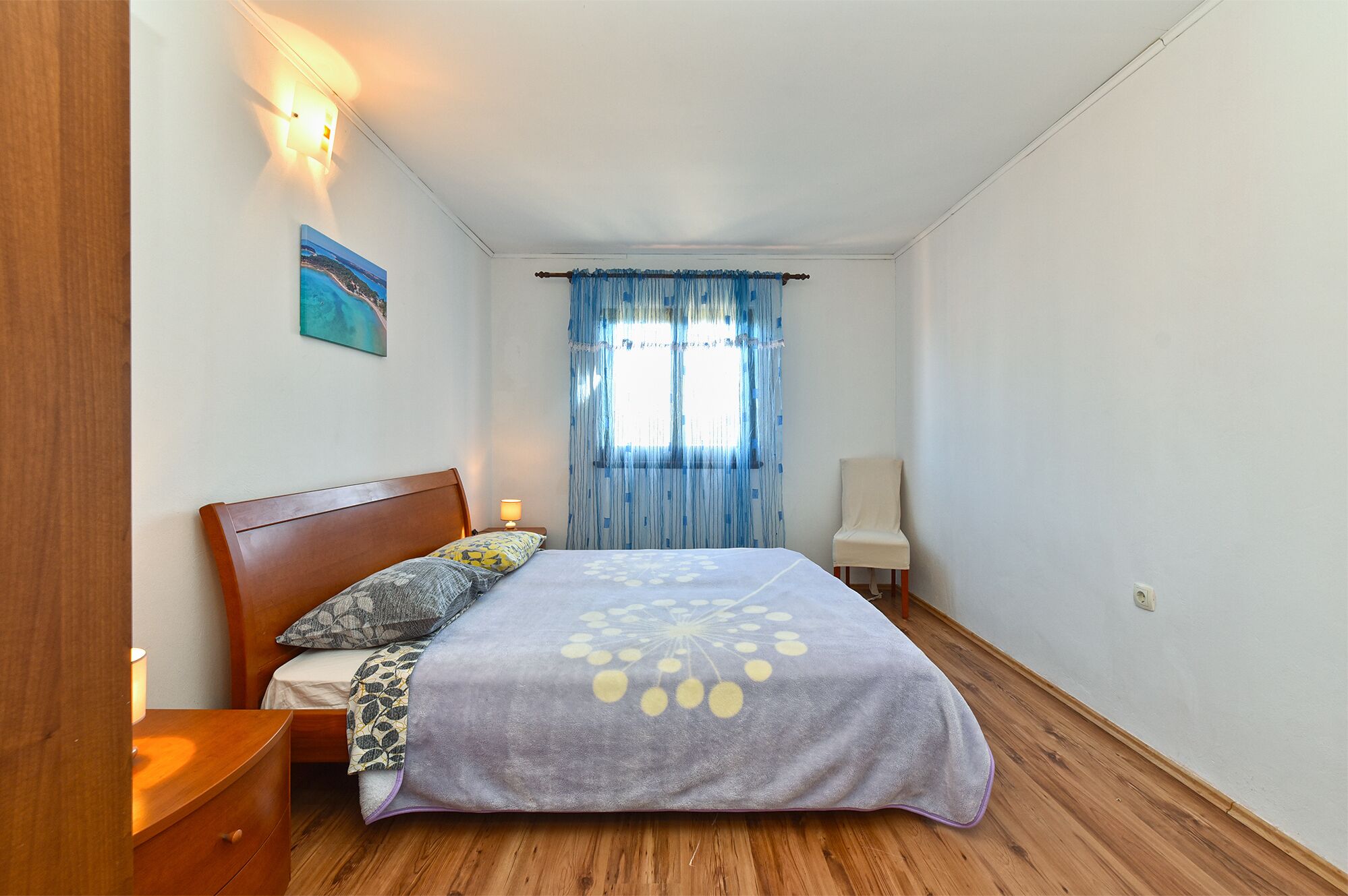 Un dormitorio con una cama de madera, una mesita de noche con lámpara, una ventana con cortinas azules y un cuadro en la pared.