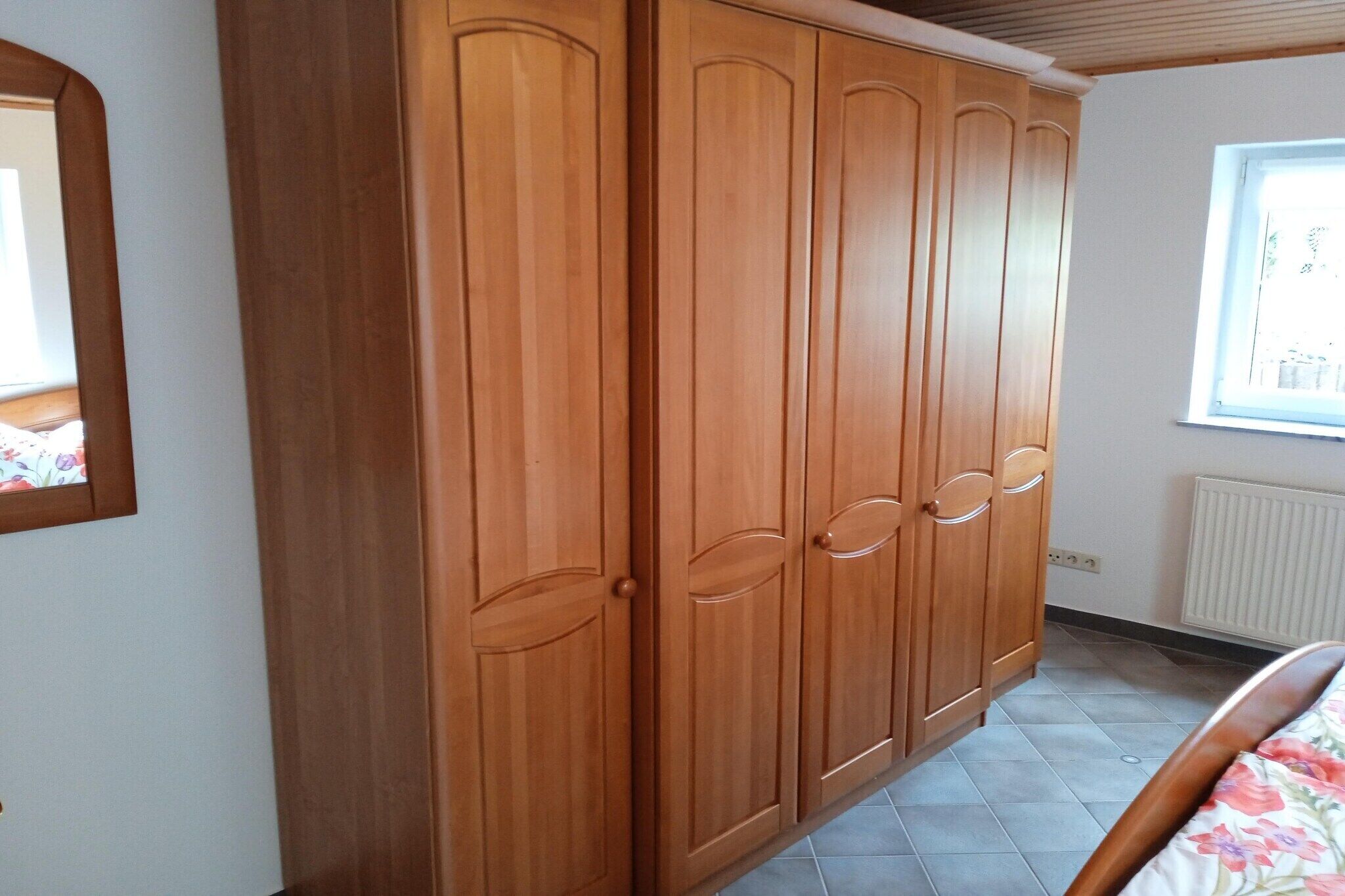A bedroom with a wooden wardrobe, a bed with a floral bedspread, a window with a view, and a radiator.