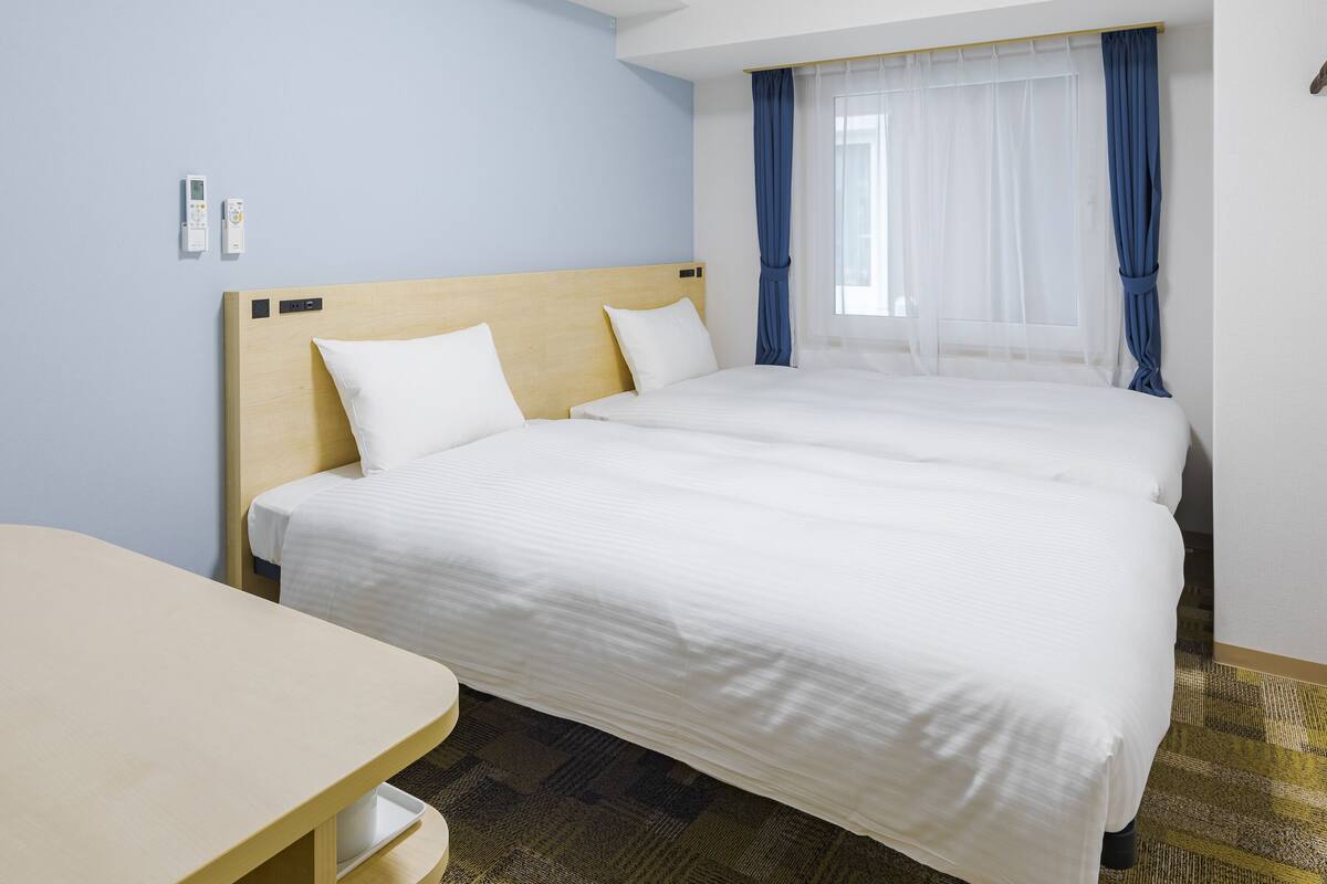 A hotel room with two beds, a wooden headboard, a desk, and a window with blue curtains.