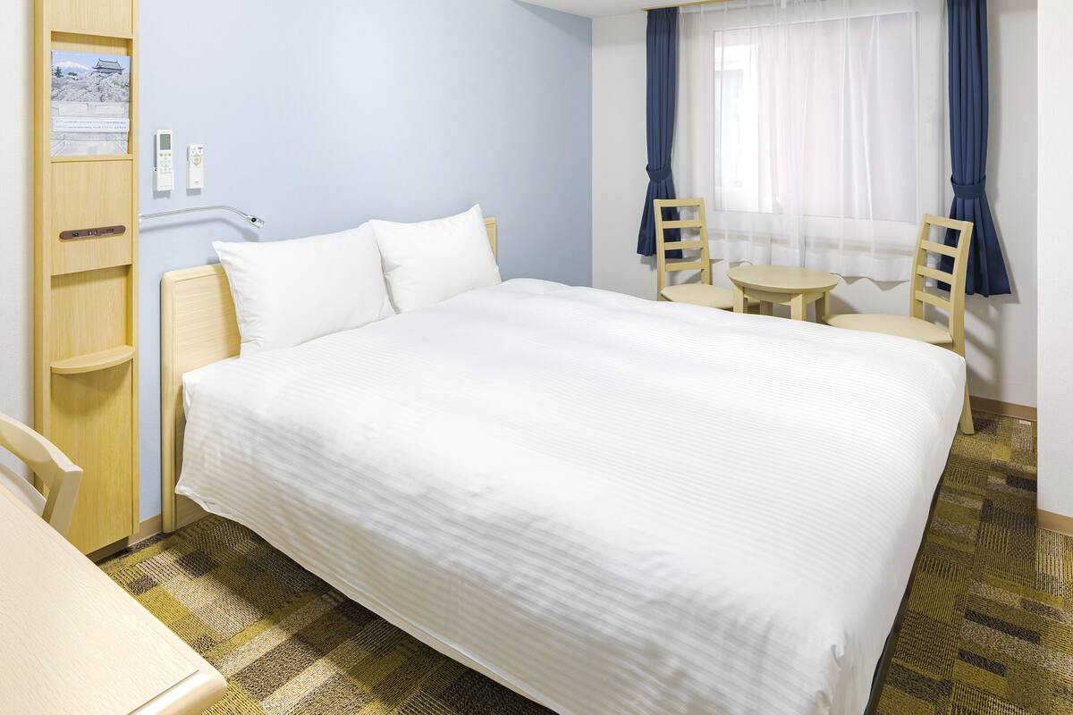 A hotel room with a large bed, a desk, a chair, a small table, and a window with curtains.