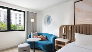Een moderne hotelkamer met een gebogen blauwe bank, een ronde witte tafel, een bed met witte lakens en uitzicht op een gebouw door een groot raam.