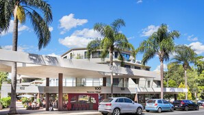 A modern building with a covered outdoor seating area, palm trees, and cars parked in front.