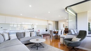 A modern living room with a grey sofa, a round coffee table, and a grey armchair. There is a dining area with a table and chairs, and a kitchen in the background.