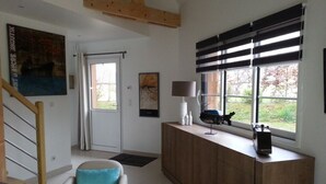 A modern living room with a wooden staircase, a large window with blinds, and two armchairs with blue cushions. - Le Clos du Héron Perché (HOUDETOT)