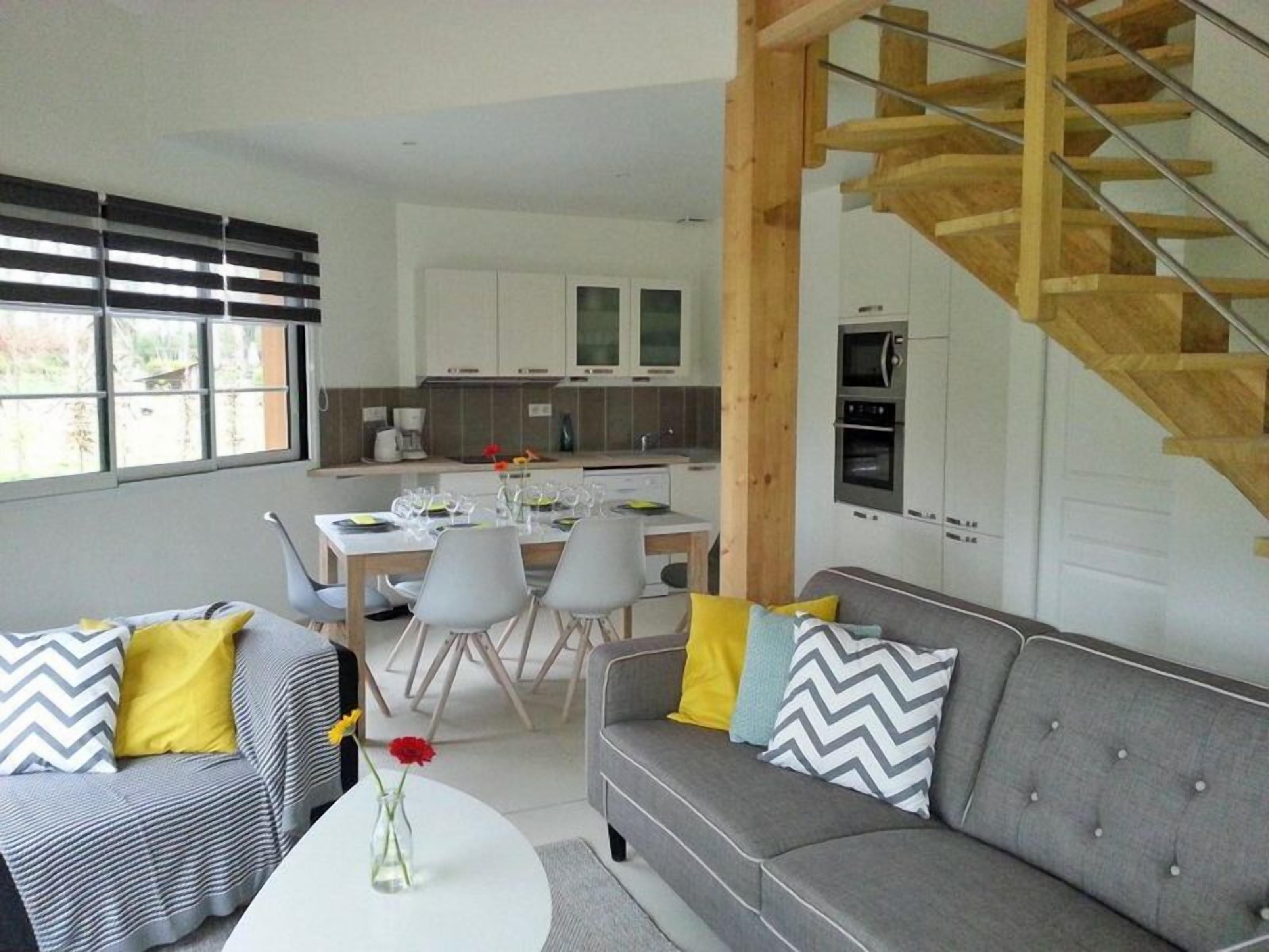 Un salon moderne avec un canapé gris, une table à manger avec des chaises, et une cuisine en arrière-plan.