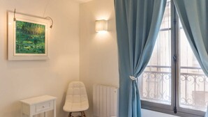 A bedroom with a bed, a white chair, a bedside table, a radiator, a window with blue curtains, and a wall-mounted light fixture. - Les Amants de Saint Jean (DIEPPE)
