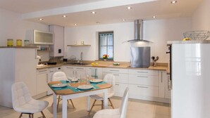 A modern kitchen with white cabinetry, a stainless steel range hood, and a dining area with white chairs and a wooden table. - Les Amants de Saint Jean (DIEPPE)