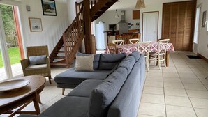 Una sala de estar con un sofá gris, una escalera de madera, un comedor con mesa y sillas, y una cocina al fondo.