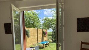 Una habitación con puerta corredera de vidrio que da a un patio trasero con mesa y sillas, cerca de una valla de madera y un jardín.