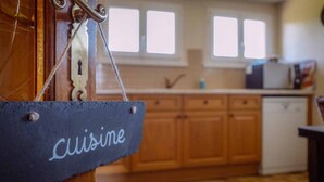 Une cuisine avec des meubles en bois, un tableau noir sur lequel est écrit le mot « Cuisine », et une fenêtre avec des stores.