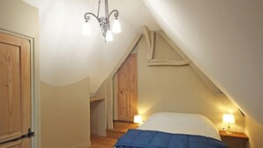 Une chambre avec un plafond en pente, une porte en bois et un lit recouvert d’une couverture bleue. On y trouve deux lampes de chevet et un lustre.