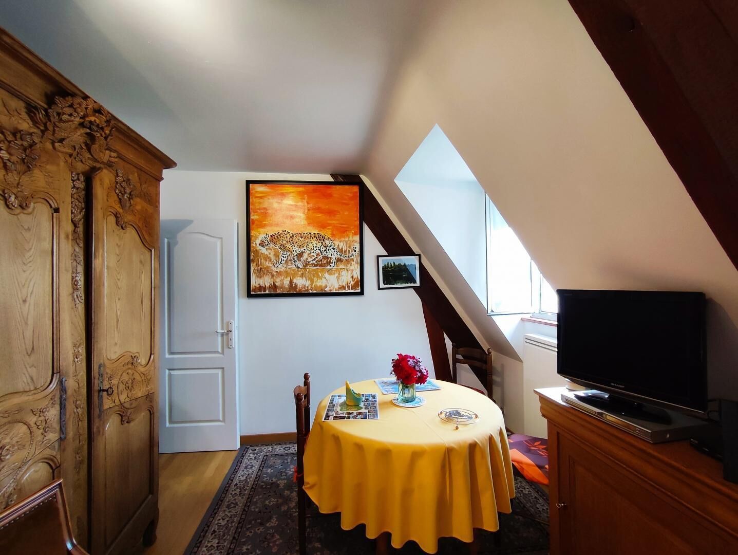 Un comedor con una mesa preparada para dos, un cuadro sobre la mesa y un televisor en un mueble a la derecha.