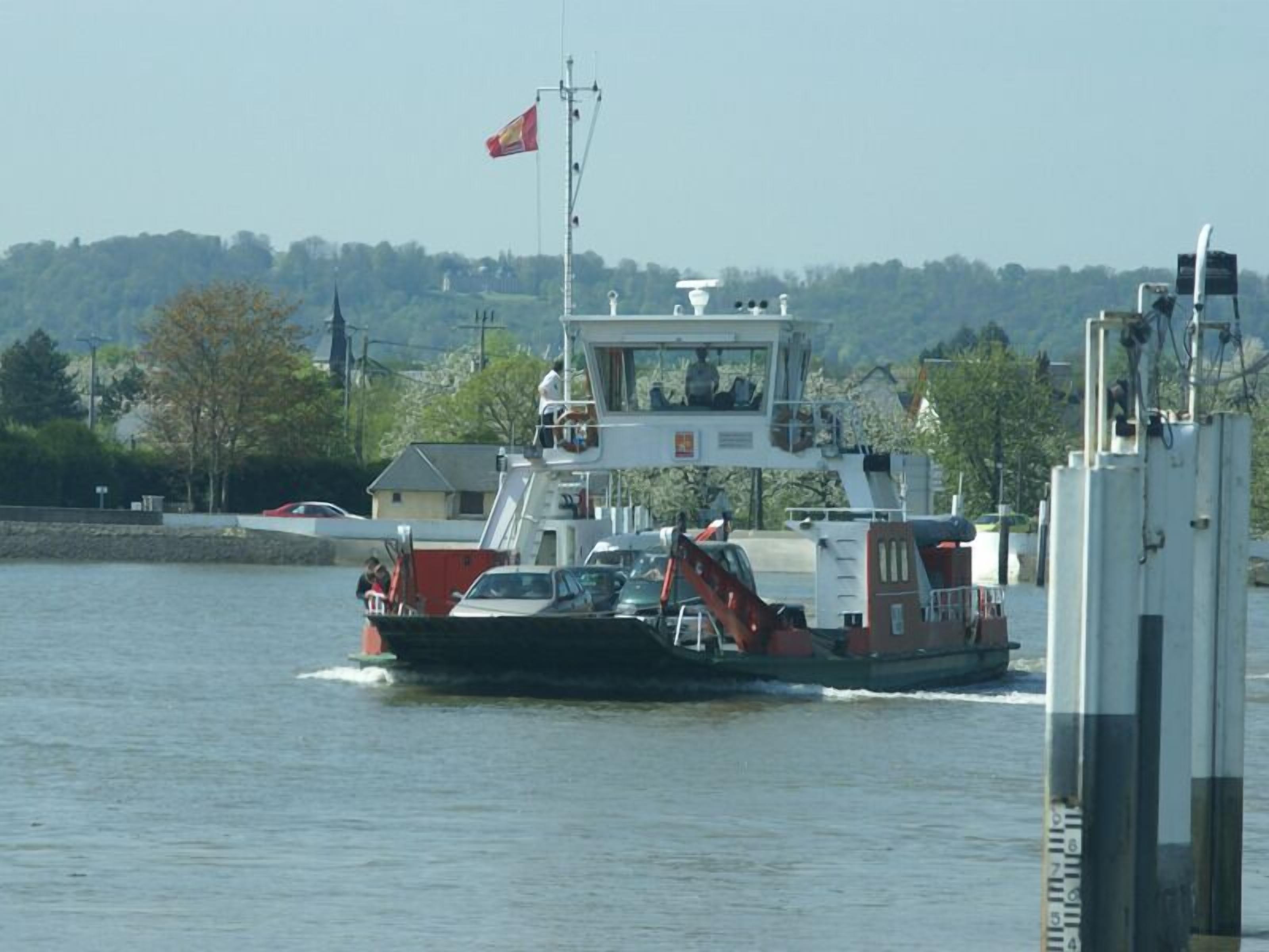 Eine Fähre mit Kabine und einer roten Flagge, die sich auf dem Wasser bewegt, im Hintergrund sind ein angedocktes Auto und Gebäude zu sehen.