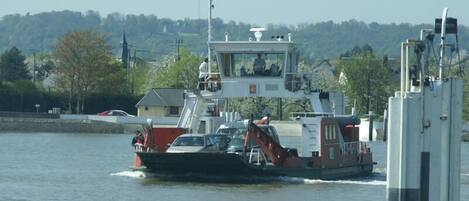Eine Fähre mit Kabine und einer roten Flagge, die sich auf dem Wasser bewegt, im Hintergrund sind ein angedocktes Auto und Gebäude zu sehen.