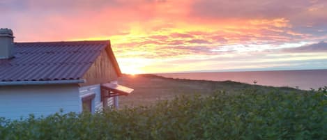 A house with a sloped roof and a porch, overlooking a body of water with a sunset in the background.