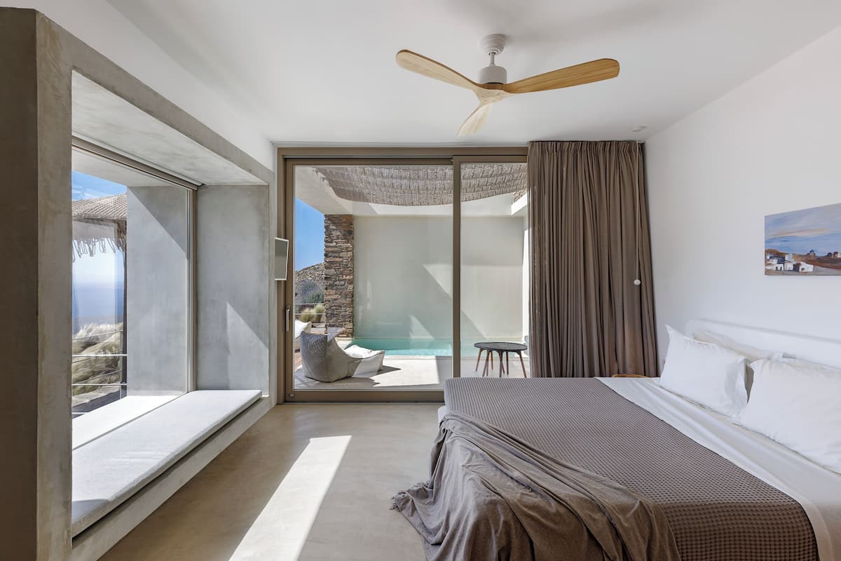 Un dormitorio con una cama grande, ventilador de techo, puerta corrediza de vidrio que da a una zona de piscina y una pared de piedra al fondo.