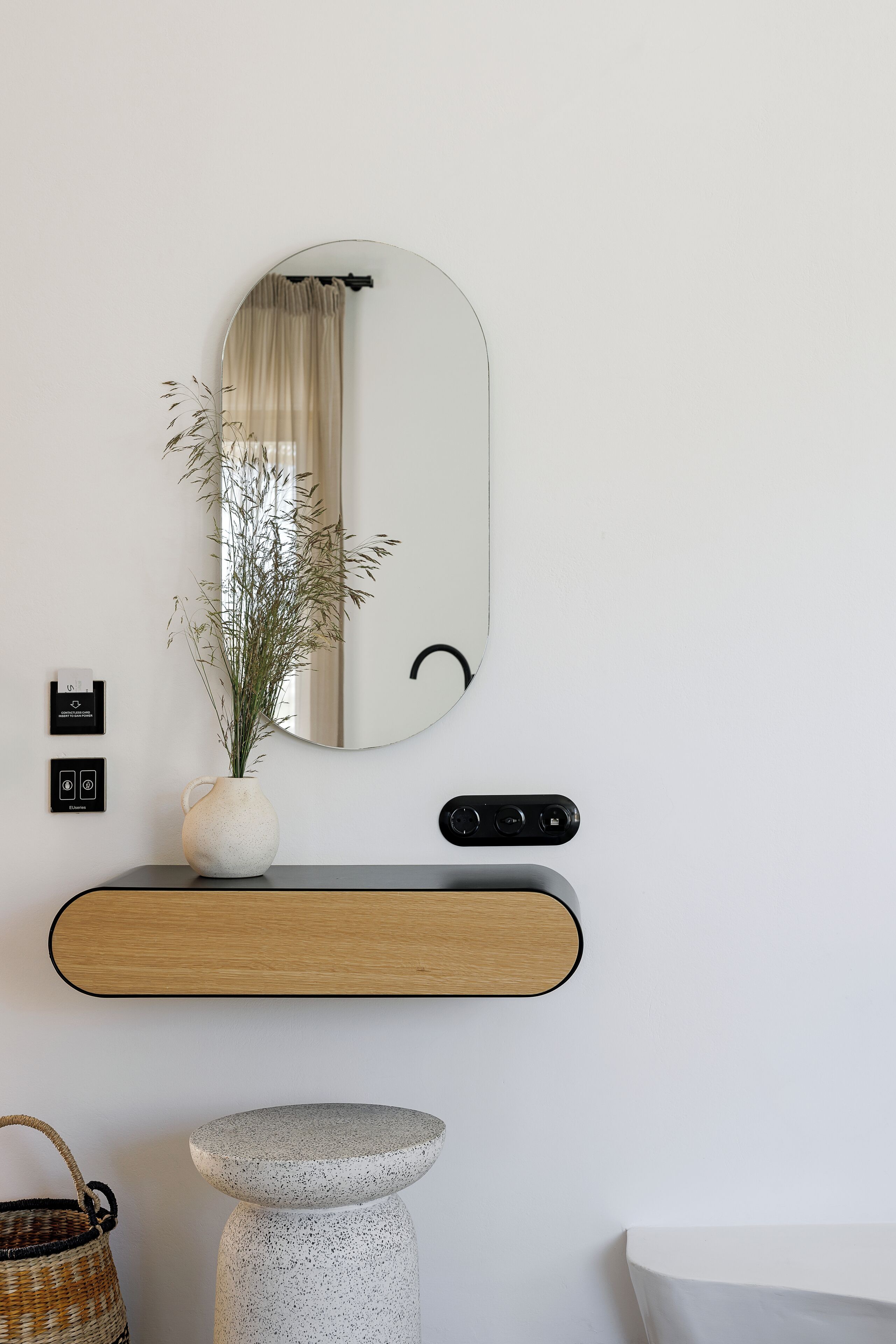 A modern bathroom with a round mirror, a vase with dried plants, a wooden console, a stone pedestal, and a wicker basket.