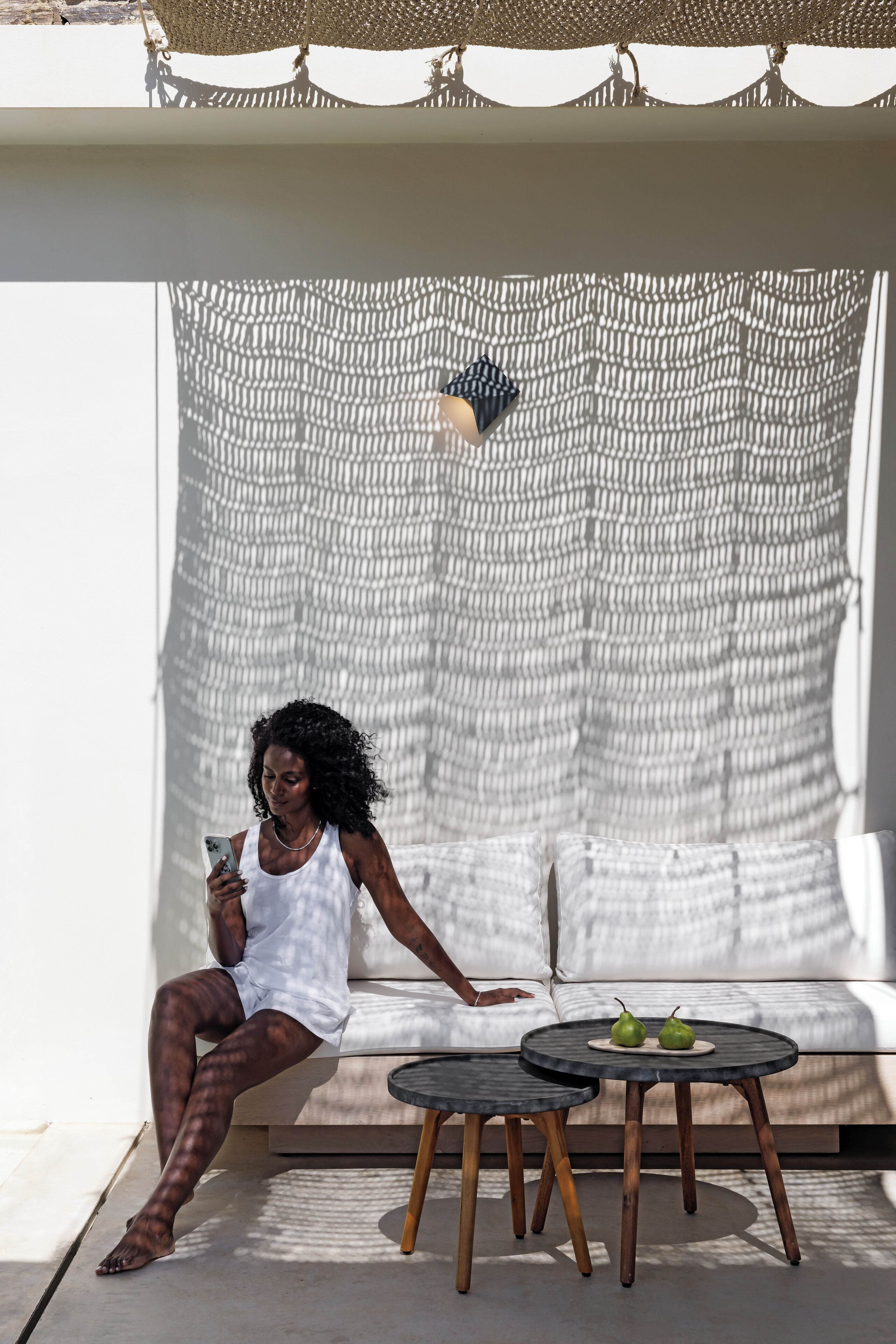 A person sitting on a white sofa with a woven net backdrop, two round tables with a plate of green pears, and a small decorative item above the net.
