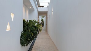 A narrow, well-lit corridor with white walls, a stone wall on one side, and a row of potted plants. - Ivy's Natural Resort (Tinos)